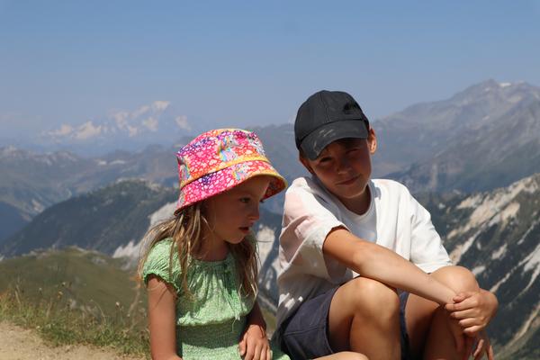 Two children are sitting on a grassy mountain ridge, with a vast expanse of mountains stretching into the distance under a clear blue sky